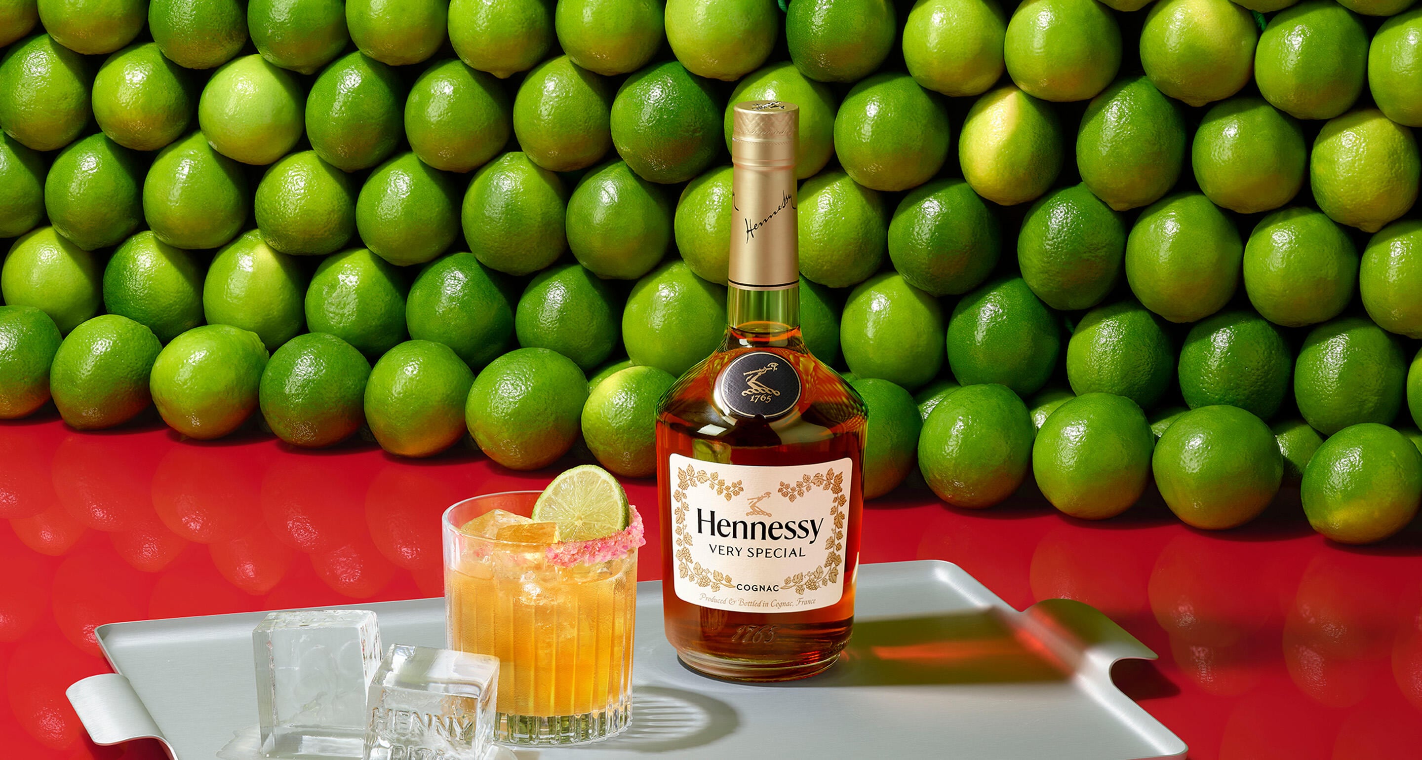 Bouteille de cognac Hennessy VS devant un mur de citrons verts, à côté d'un verre de margarita au cognac