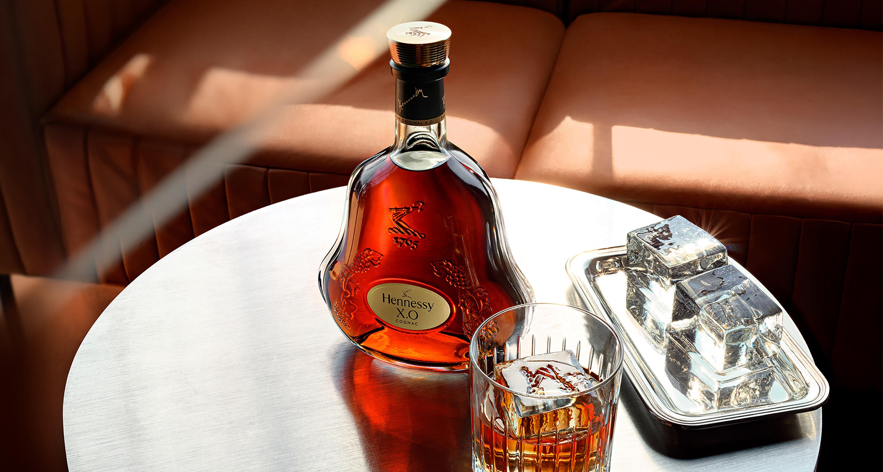 Carafe de cognac Hennessy X.O sur une table basse, à côté d'un verre de cognac servi avec un glaçon