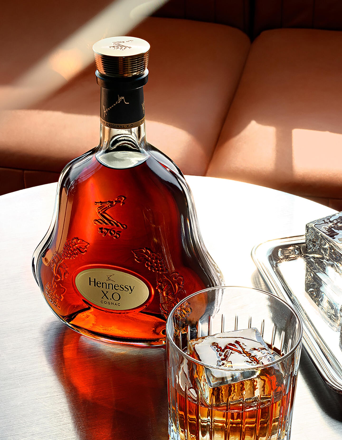 Carafe de cognac Hennessy X.O sur une table basse, à côté d'un verre de cognac servi avec un glaçon