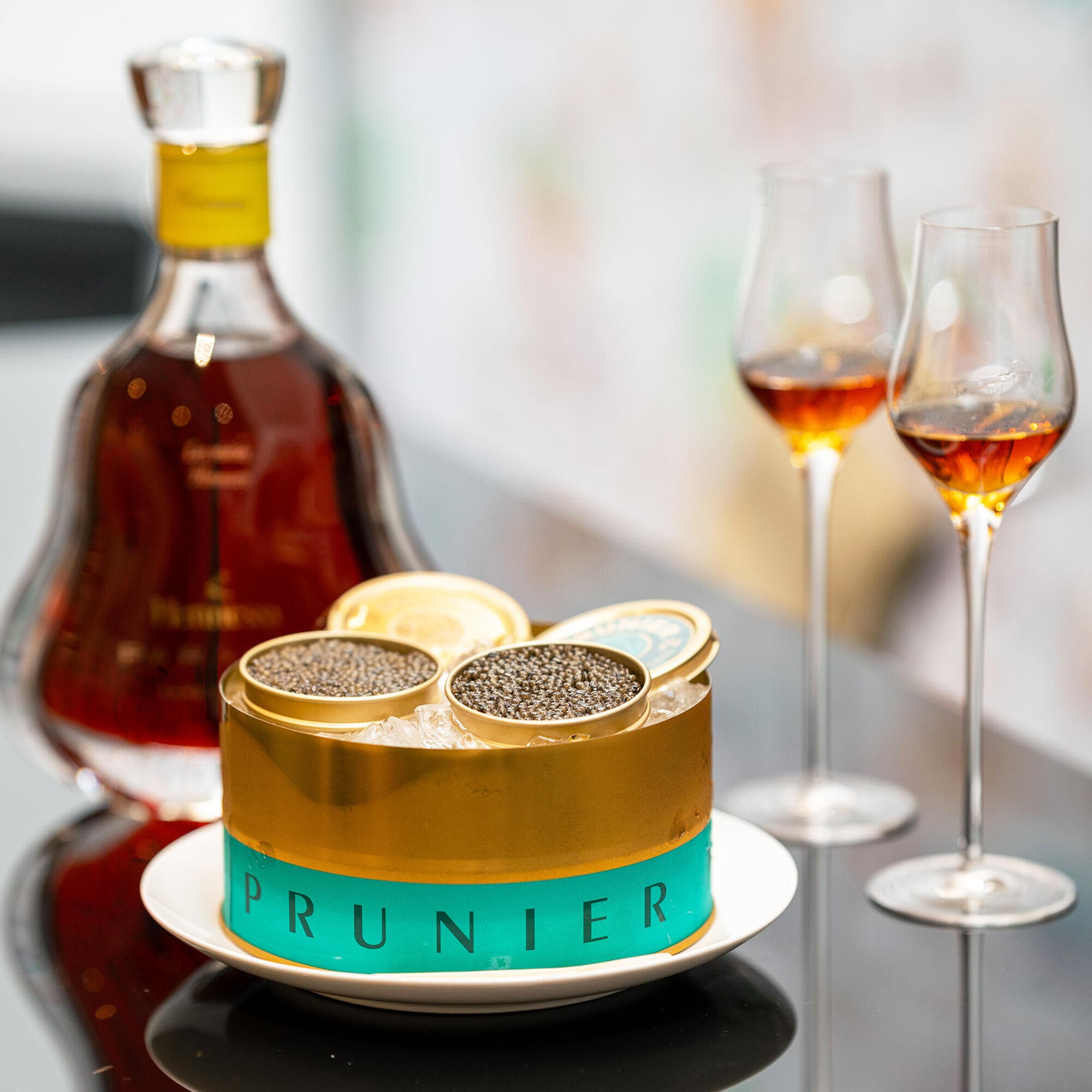 Carafe de cognac Hennessy Paradis à côté d'une boite de caviar Prunier turquoise et dorée, avec 2 verres de cognac
