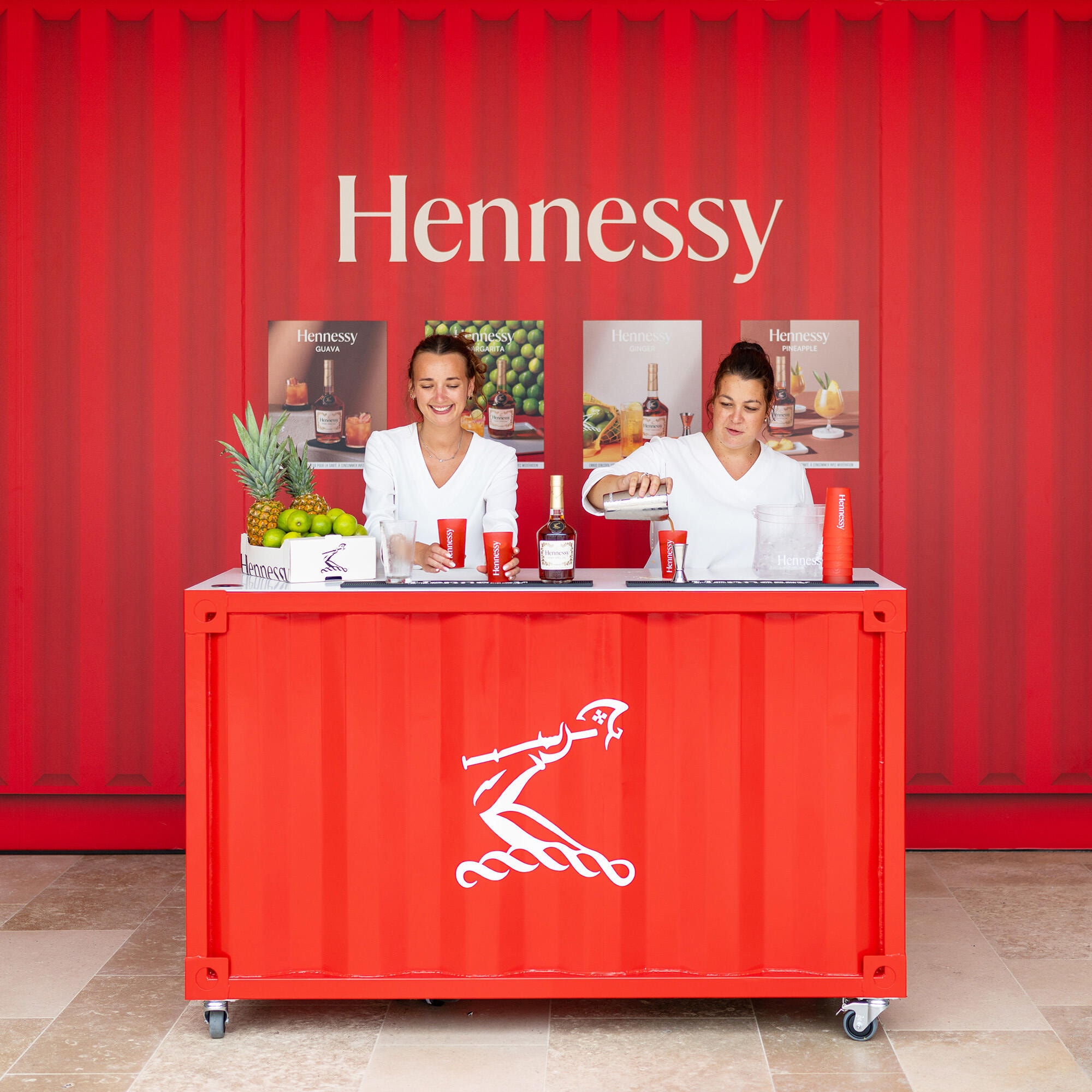 Jeanne et Céline, bartenders de la Maison Hennessy, devant un bar et un mur rouge, servant des cocktails au cognac sur la terrasse éphémère de la Boutique Hennessy à Cognac
