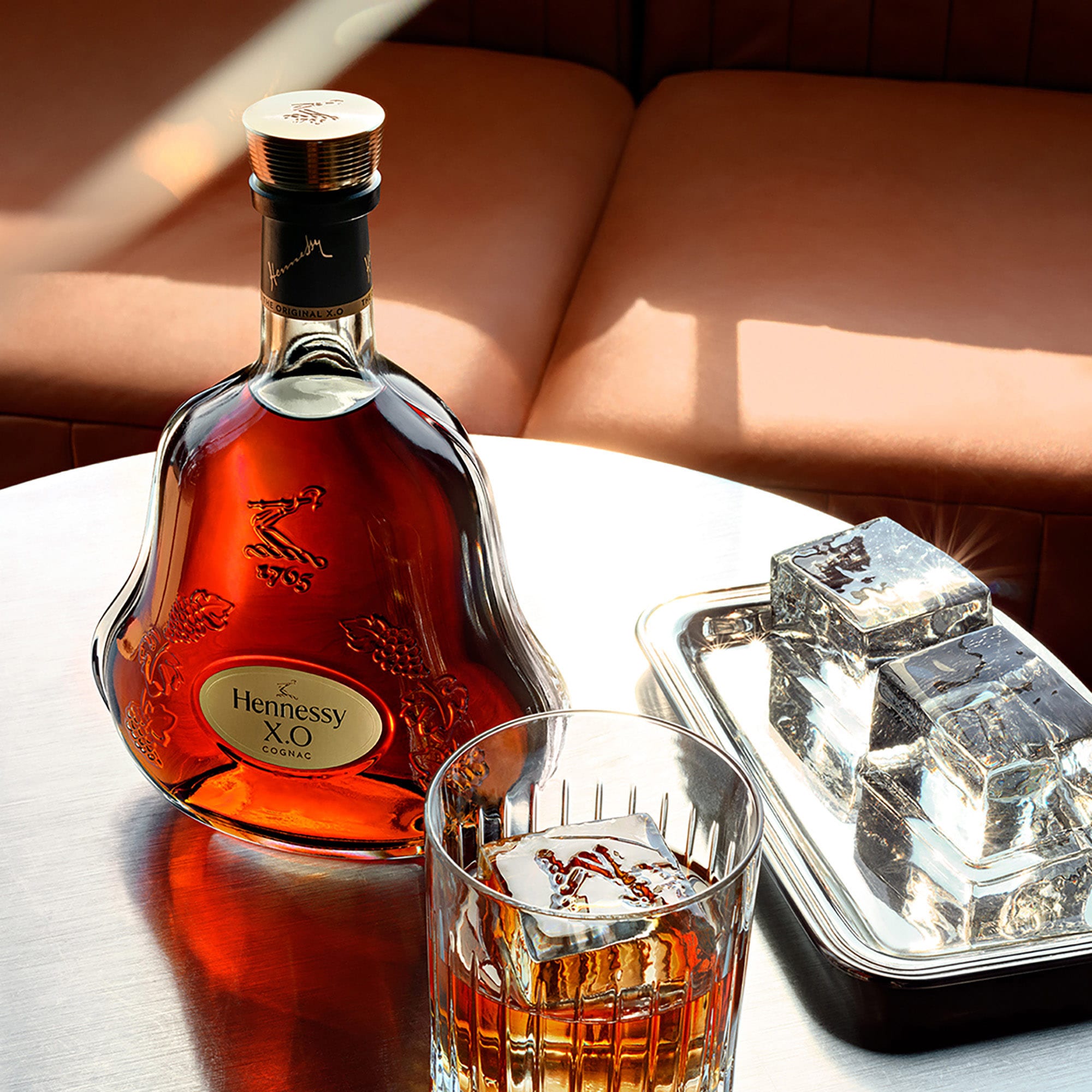 Carafe de cognac Hennessy X.O sur une table basse, à côté d'un verre de cognac servi avec un glaçon