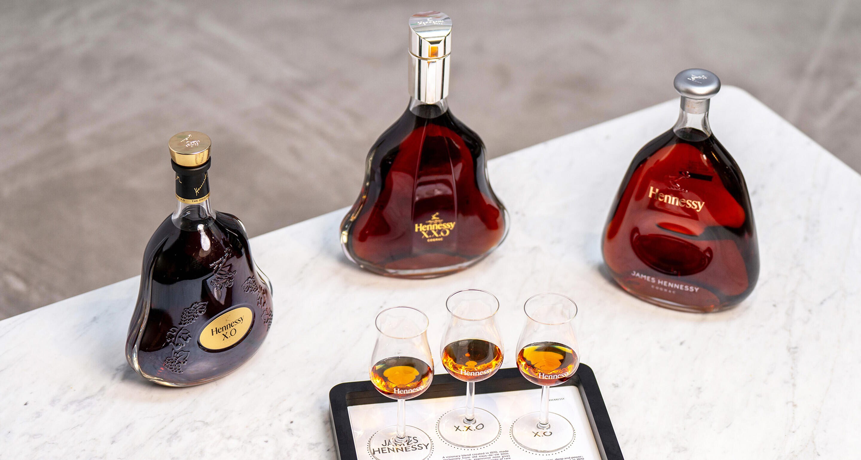 Decanters of James Hennessy, Hennessy XO and Hennessy XXO on a coffee table, in front of 3 matching glasses of cognac